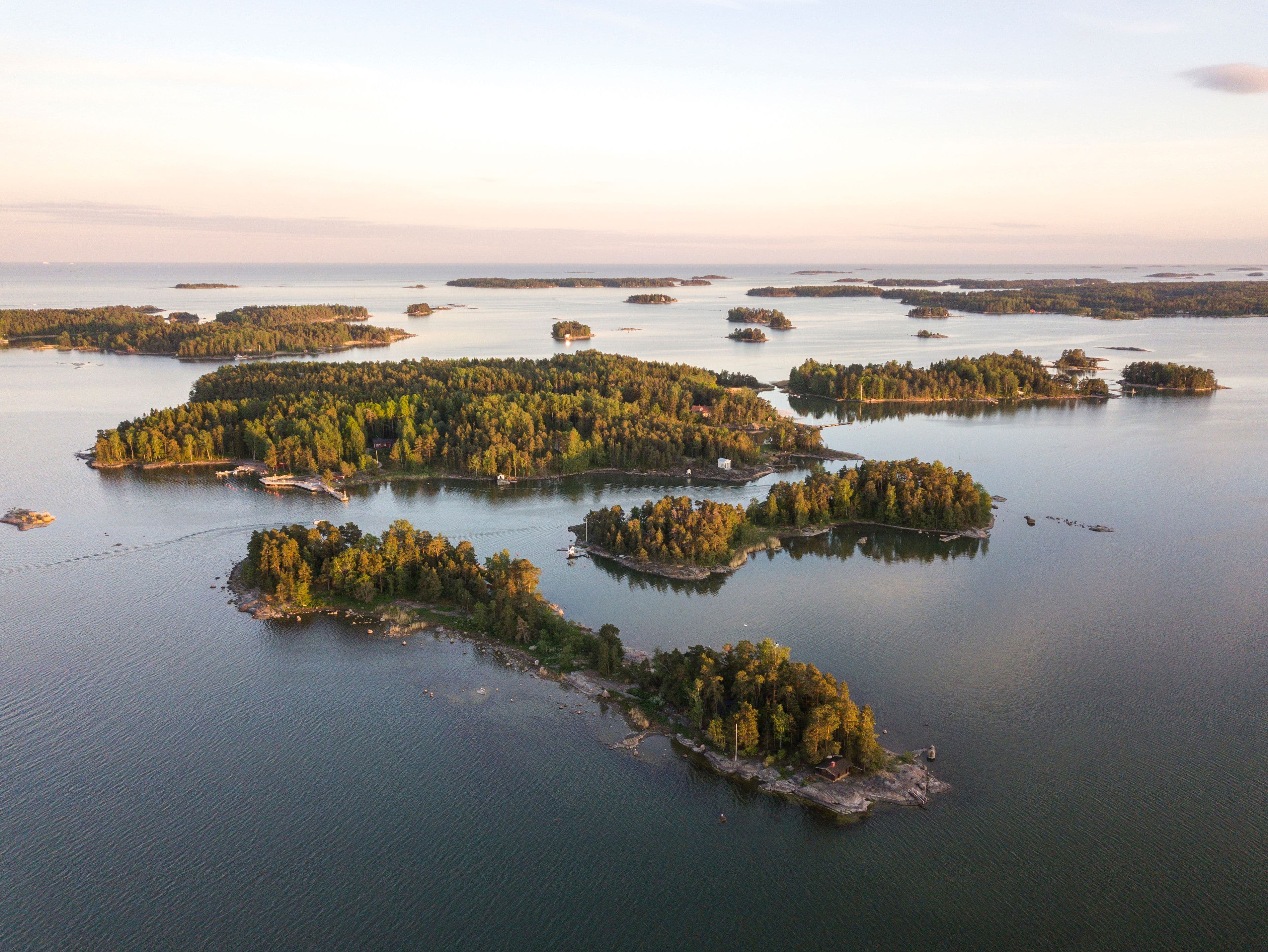 Ilmakuva Espoon saaristosta, lukuisia metsäisiä saaria ja salmia, tyynessä meressä auringonlaskun aikaan