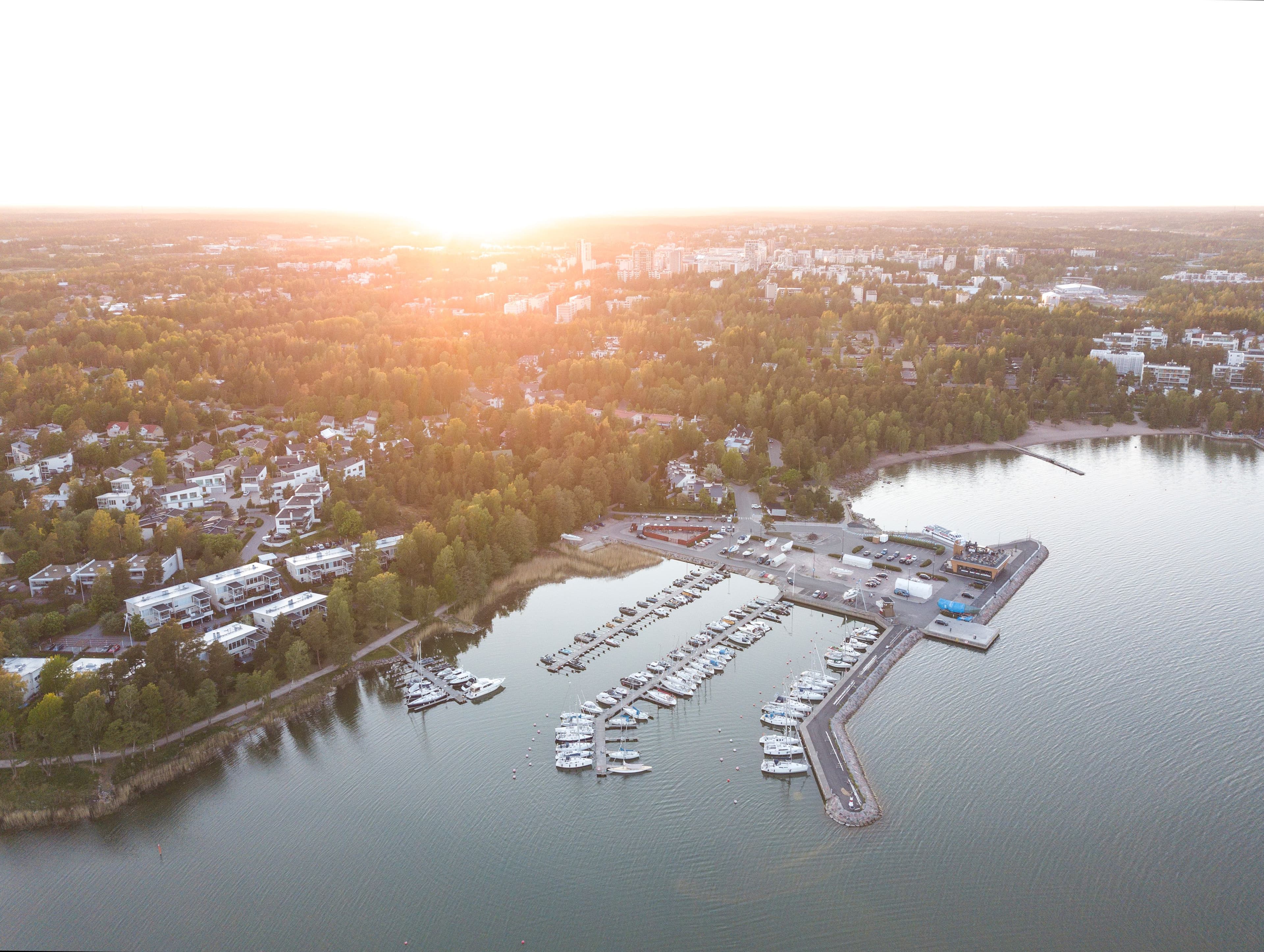 Ilmakuva Helsingin Lauttasaaresta auringonlaskun aikaan, etualalla venesatama täynnä veneitä, taustalla asuinalue ja kaupungin siluetti